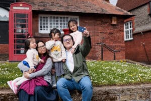 A family of two parents and three young children are taking a selfie outside the Tudor Rose Tea Rooms.